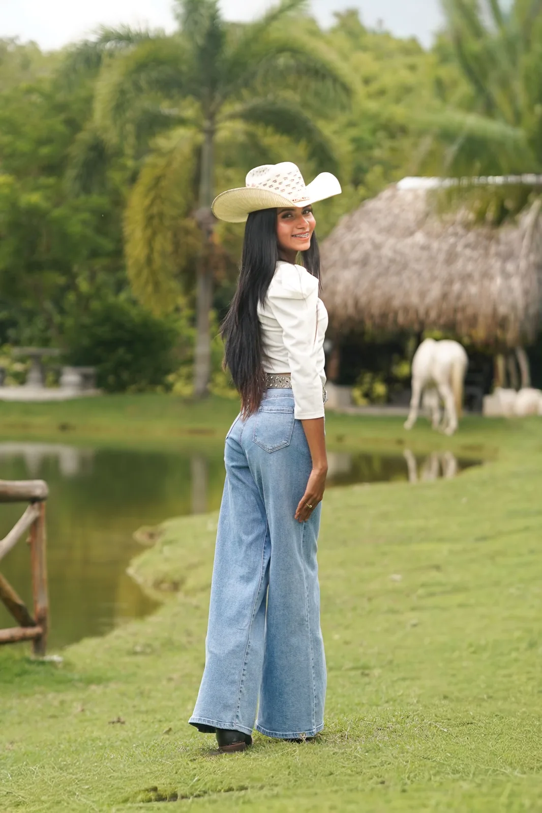 Joven con sombrero vaquero junto a un estanque, con un caballo blanco al fondo