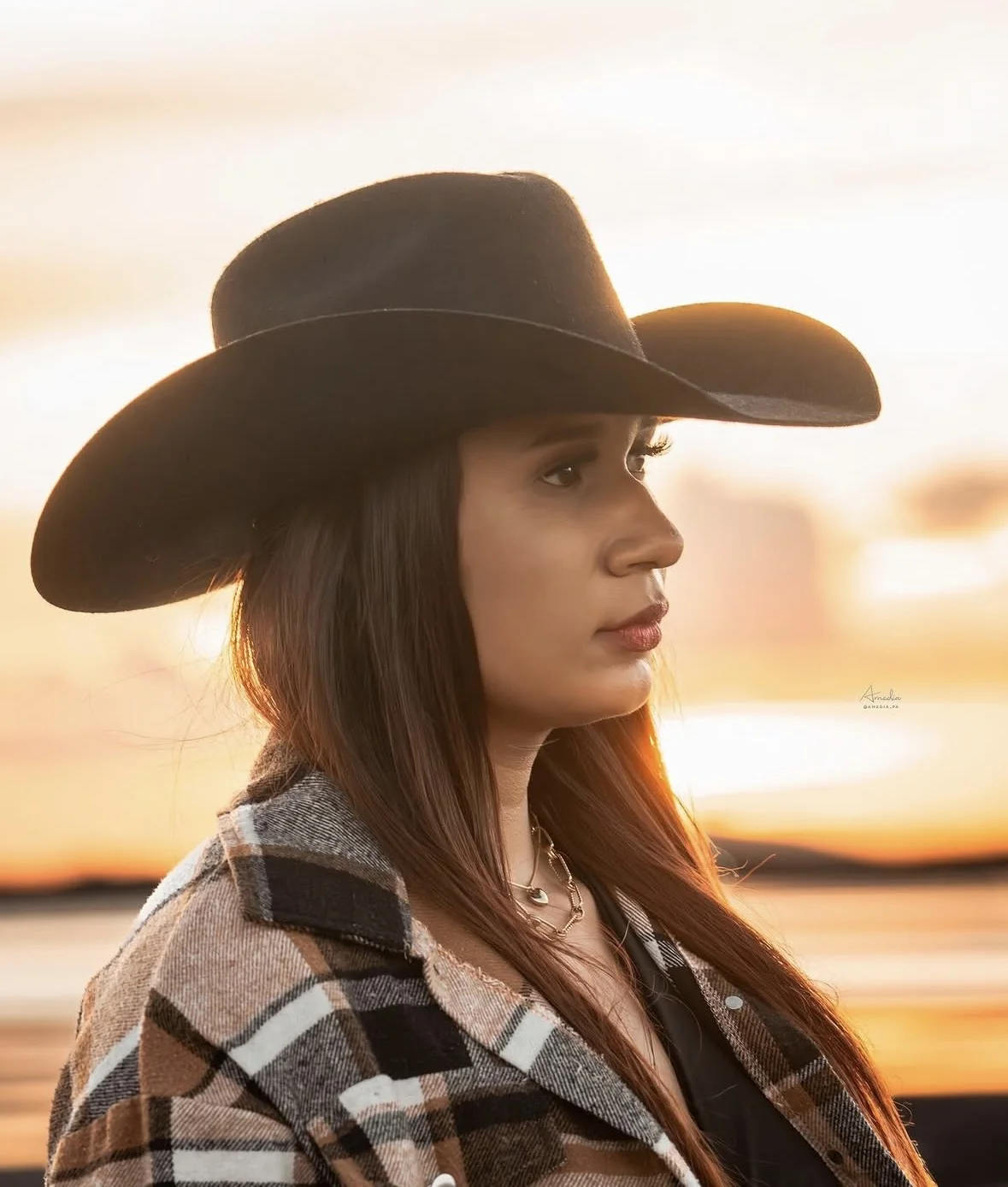 Joven de perfil con sombrero vaquero y camisa a cuadros frente al atardecer