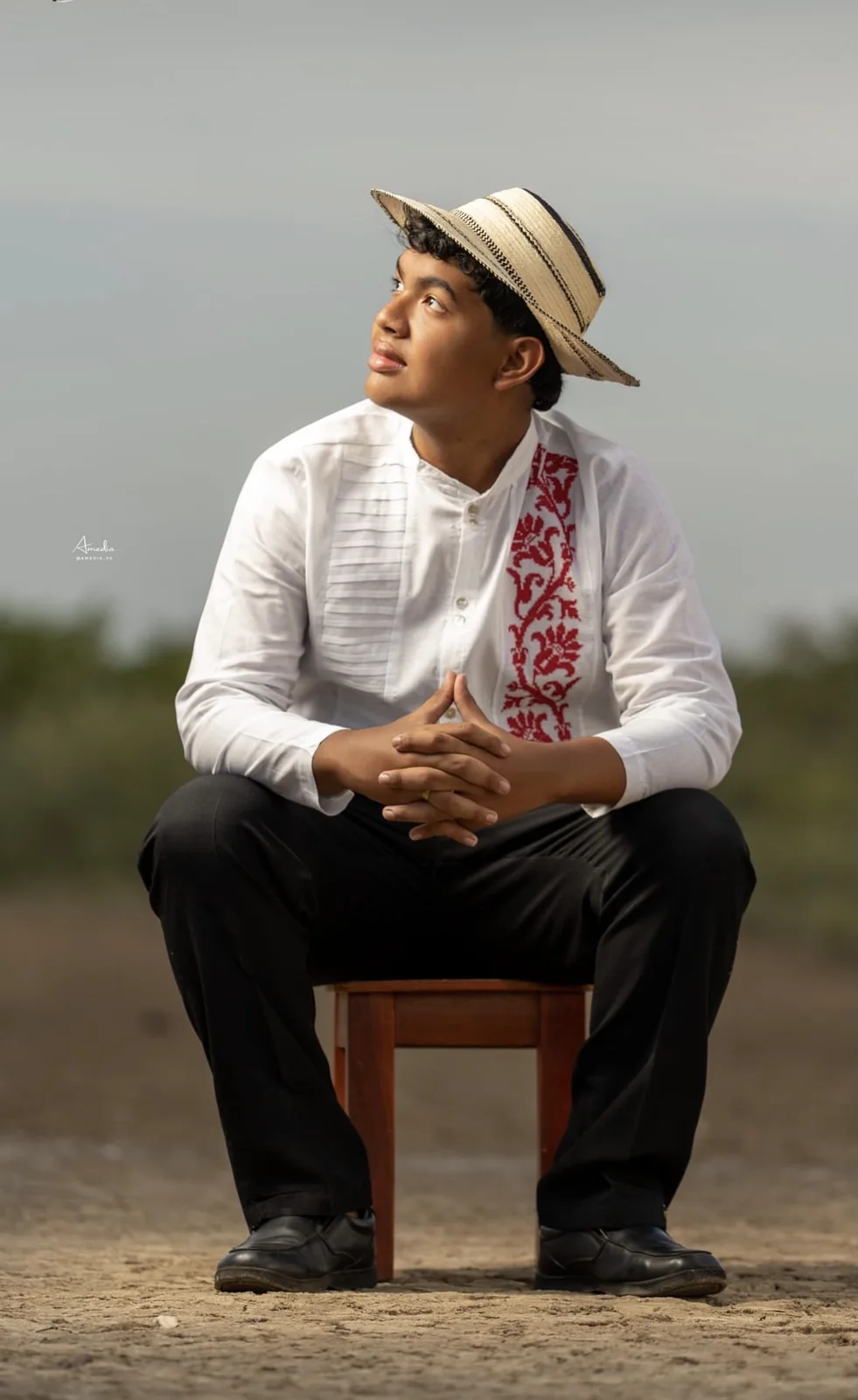 Joven con camisa montuna bordada y sombrero pintao, sentado en un taburete